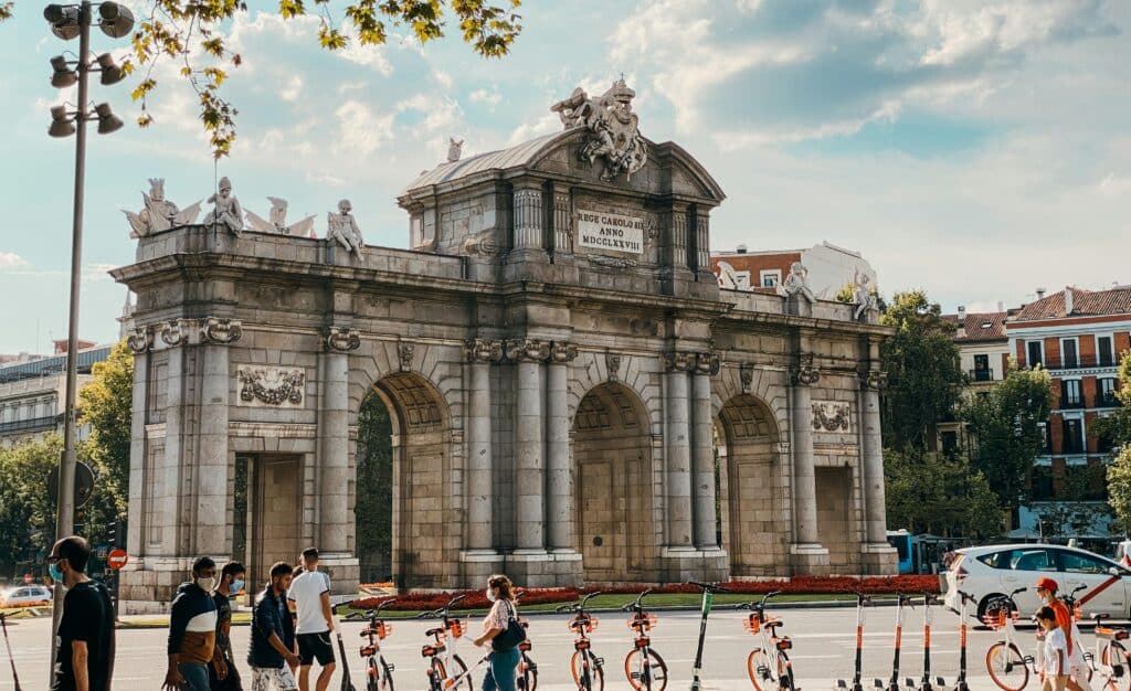 pexels-photo-16121318-16121318 View of Puerta de Alcalá with pedestrians and scooters in vibrant Madrid.